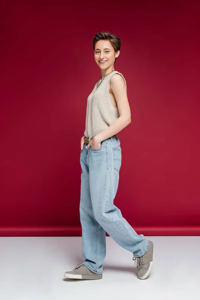 Piena lunghezza della giovane donna allegra con i capelli corti che cammina con le mani in tasche di jeans su sfondo rosso scuro — Foto stock