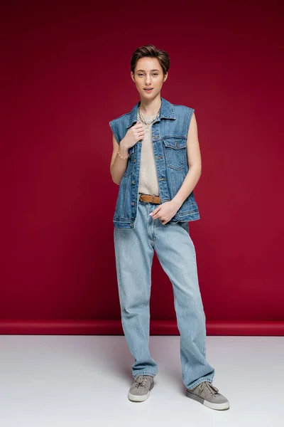 Comprimento total da jovem elegante com cabelo curto posando em roupa jeans no fundo vermelho escuro — Fotografia de Stock