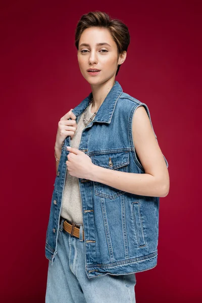 Stylische junge Frau mit kurzen Haaren posiert in Jeansweste auf dunkelrotem Hintergrund — Stockfoto