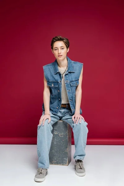 Полная длина стильная молодая женщина с короткими волосами сидя в джинсовой одежде на темно-красном фоне — стоковое фото