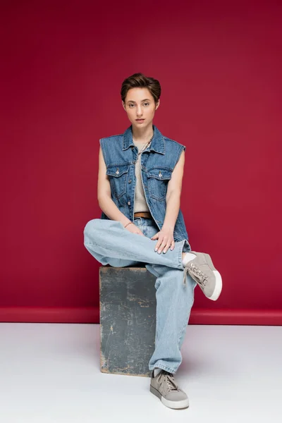 Full length of stylish model with short hair sitting in denim outfit on dark red background — Stock Photo