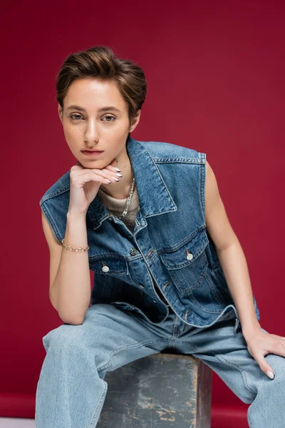 Стильна молода модель з коротким волоссям, що сидить у джинсовому вбранні на темно-червоному тлі — стокове фото
