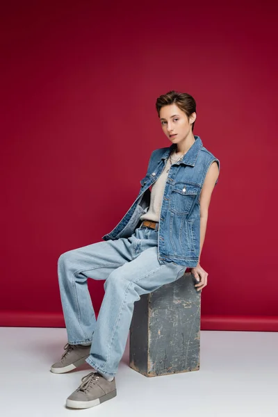 Полная длина симпатичной молодой модели с короткими волосами сидя в джинсовой одежде на темно-красном фоне — стоковое фото