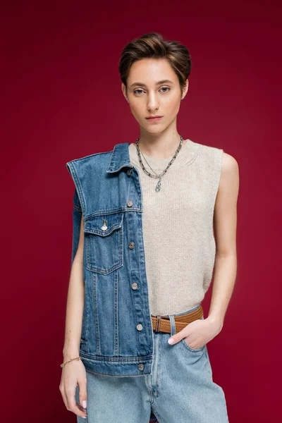 Красива молода модель в джинсовому жилеті, що стоїть з рукою в кишені на темно-червоному тлі — стокове фото