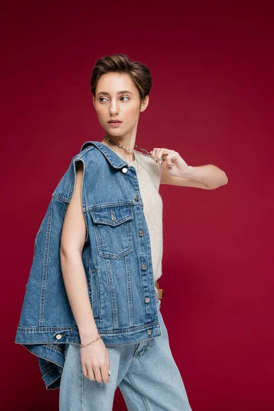 Довольно молодая модель в джинсовой жилетке тянет цепочку ожерелья, позируя на темно-красном фоне — стоковое фото
