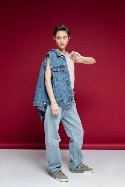 Piena lunghezza di bella giovane donna in gilet di jeans tirando catena collana mentre posa su sfondo rosso scuro — Foto stock