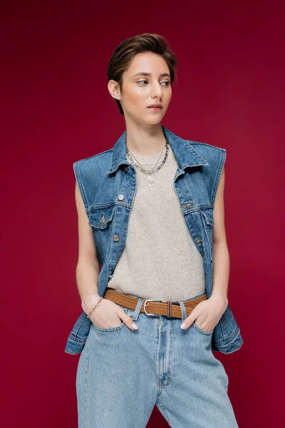 Молода модель в джинсовому жилеті, що стоїть руками в кишенях на темно-червоному тлі — стокове фото