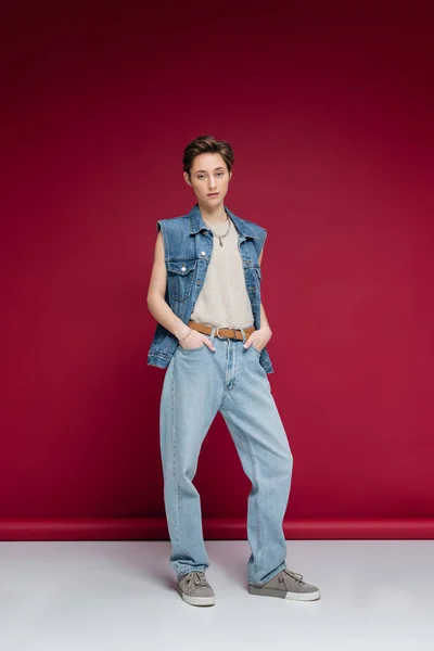 Повна довжина молодої моделі в джинсовому вбранні, що стоїть руками в кишенях на темно-червоному тлі — Stock Photo