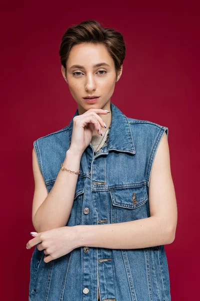 Молодая модель с короткими волосами позирует в джинсовой жилетке на темно-красном фоне — стоковое фото