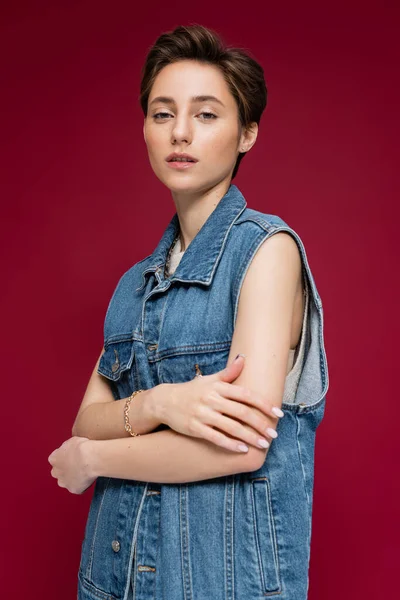 Jeune modèle avec les cheveux courts regardant la caméra tout en posant sur fond rouge foncé — Photo de stock