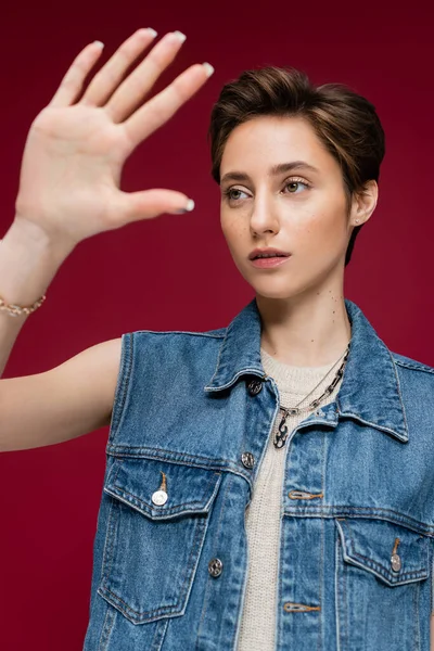 Young model in denim vest standing with outstretched hand on maroon background — Stock Photo