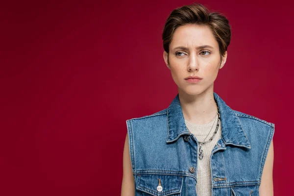 Suspicious young woman in denim vest looking away on burgundy background — Stock Photo