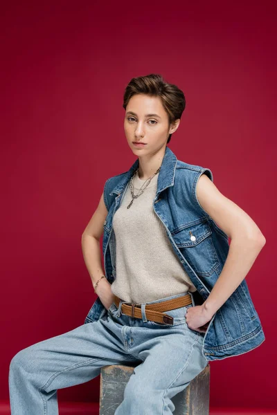 Stylish model in denim outfit with vest posing with hands on hips on burgundy background — Stock Photo
