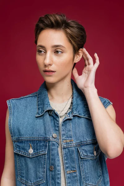Stylish model in denim outfit with vest fixing her short hair on burgundy background — Stock Photo