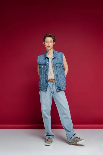 Повна довжина стильної моделі в джинсовому вбранні з жилетом, що стоїть на бордовому фоні — стокове фото