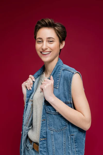 Радостная модель в джинсовой одежде с жилетом, позирующая на бордовом фоне — стоковое фото