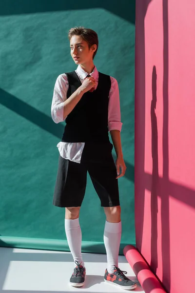 Piena lunghezza di elegante giovane donna con i capelli corti in posa in uniforme scolastica su rosa e verde — Stock Photo