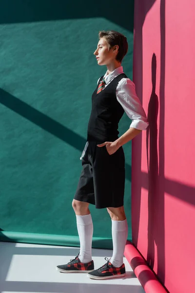 Повна довжина стильного молодого студента з коротким волоссям позує рукою в кишені чорних шортів на рожевому і зеленому — стокове фото