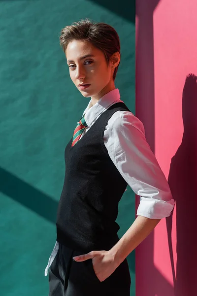 Elegante giovane studente con i capelli corti in posa con mano in tasca di pantaloncini neri su rosa e verde — Foto stock