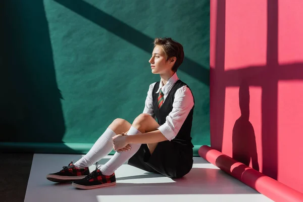 Full length of stylish young woman with short hair sitting in school uniform on green and pink background — Stock Photo