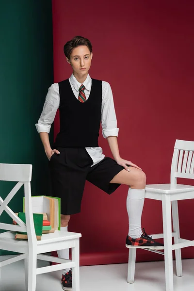 Stylish young woman with short hair posing in school uniform around chairs and books on green and pink background — Stock Photo