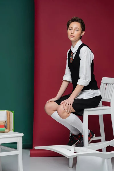 Piena lunghezza di elegante giovane studente con i capelli corti seduto sulla sedia vicino a libri su sfondo verde e rosso — Foto stock