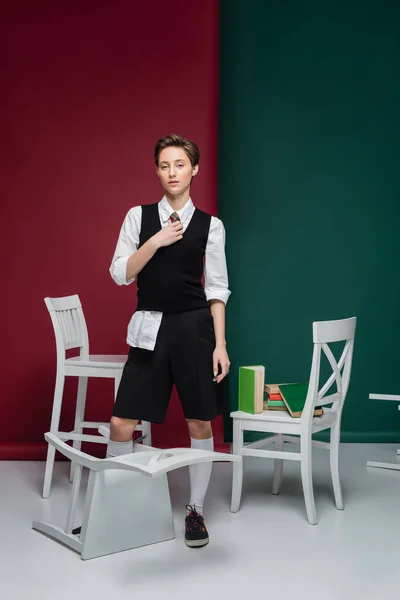 Volle Länge der stilvolle Student mit kurzen Haaren hält Buch und steht in der Nähe von Stühlen auf grünem und rotem Hintergrund — Stockfoto