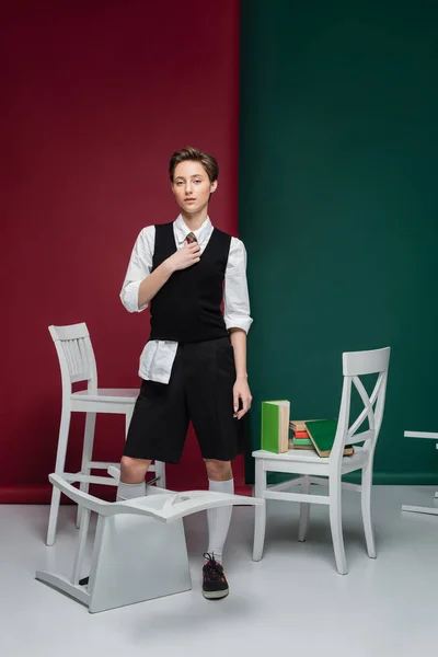 Full length of stylish student with short hair holding book and standing near chairs on green and red background — Stock Photo