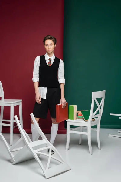 Comprimento total da jovem elegante com cabelo curto segurando livro e de pé perto de cadeiras no fundo verde e vermelho — Fotografia de Stock