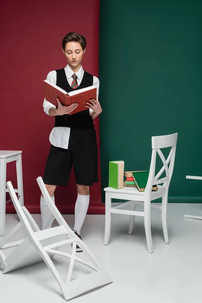 Comprimento total da jovem elegante com cabelo curto em pé perto de cadeiras e livro de leitura no fundo verde e vermelho — Fotografia de Stock