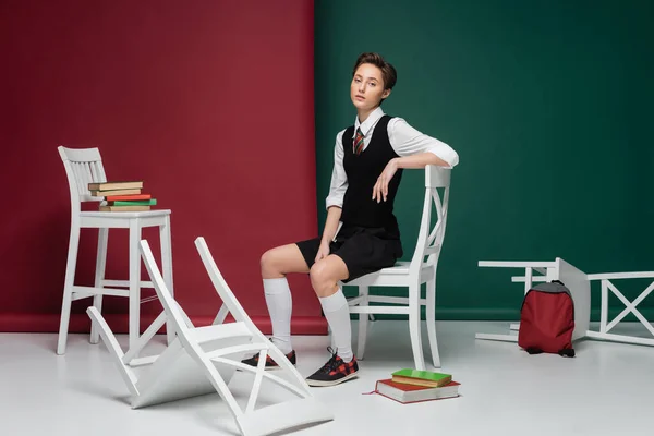 Volle Länge der stilvollen jungen Frau mit kurzen Haaren sitzt auf weißem Stuhl zwischen Büchern auf grünem und rotem Hintergrund — Stockfoto