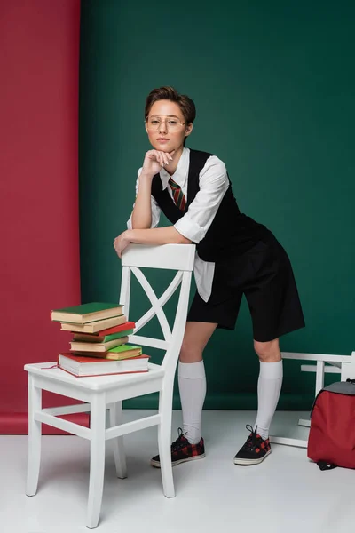 Volle Länge der stilvollen jungen Frau mit kurzen Haaren lehnt auf Stuhl mit Büchern auf grünem und rosa Hintergrund — Stockfoto