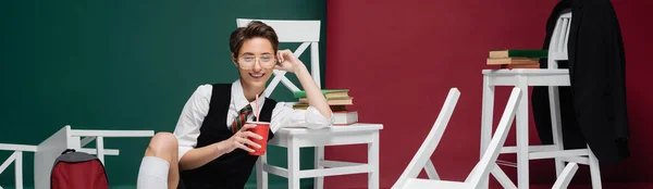 Feliz joven estudiante en anteojos celebración de la bebida en la taza de plástico cerca de sillas y libros sobre fondo verde y borgoña, pancarta - foto de stock