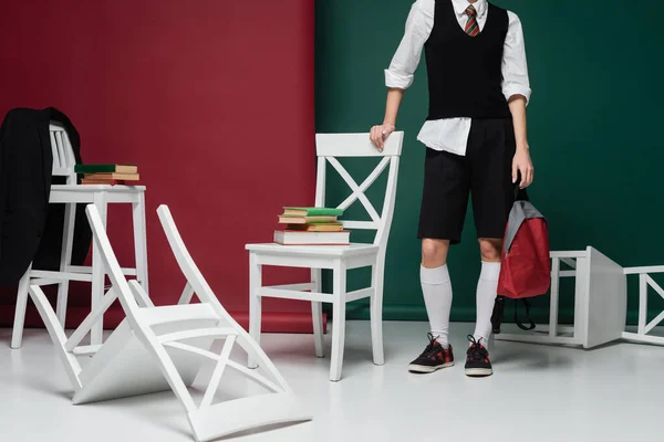 Vue recadrée du jeune étudiant élégant en uniforme scolaire debout autour des chaises avec des livres sur fond vert et bordeaux — Photo de stock