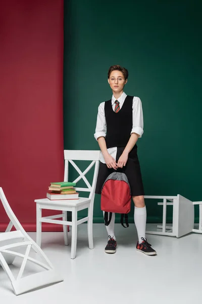 Полная длина стильный молодой студент в очках, стоящих с рюкзаком вокруг стульев и книг на зеленом и розовом фоне — стоковое фото