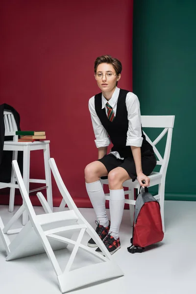 Volle Länge der stilvolle Student in Brille sitzt mit Rucksack um Stühle und Bücher auf grünem und kastanienbraunem Hintergrund — Stockfoto