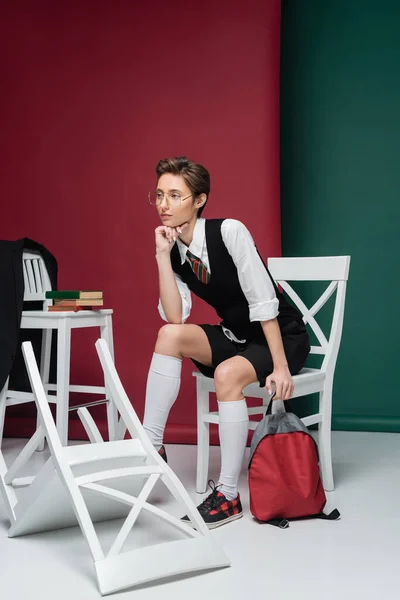 Comprimento total de jovem elegante em óculos sentados com mochila em torno de cadeiras e livros sobre fundo verde e marrom — Fotografia de Stock