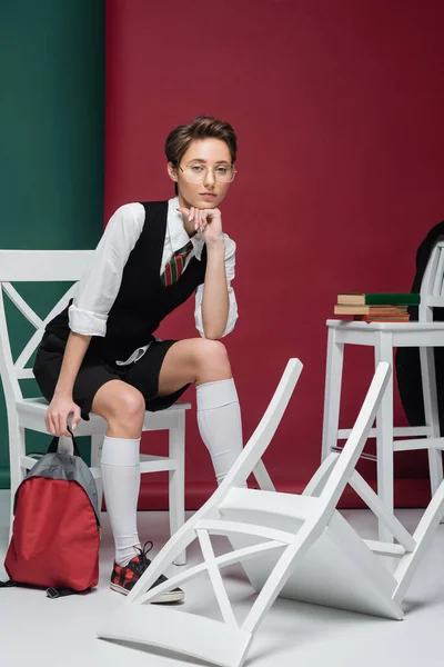 Junge Studentin mit Brille sitzt mit Rucksack auf Stühlen und Büchern auf grünem und kastanienbraunem Hintergrund — Stockfoto
