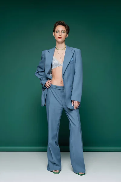 Full length of stylish woman in satin bra and blue pantsuit posing with hand on hip on turquoise green background — Stock Photo