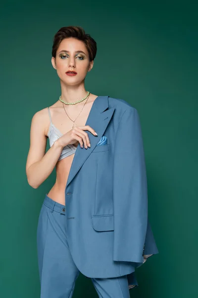 Young woman with short hair posing in satin bra and holding blue blazer near shoulder on turquoise green background — Stock Photo