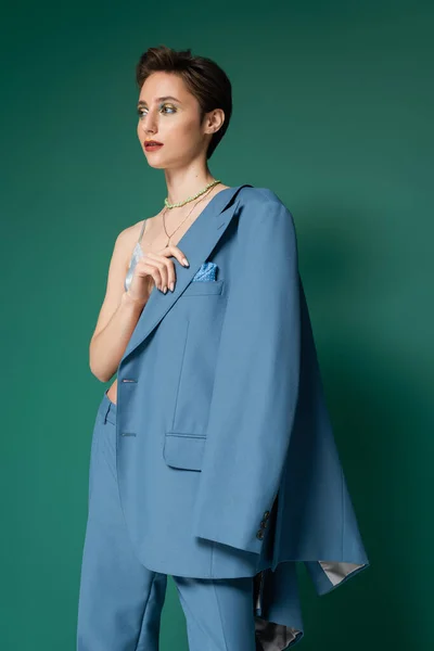 Young model with short hair posing in satin bra and holding blue blazer near shoulder on turquoise green background — Stock Photo