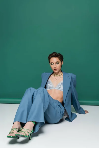 Full length of young woman with short hair posing in blue suit with satin bra and heeled sandals on turquoise green — Stock Photo