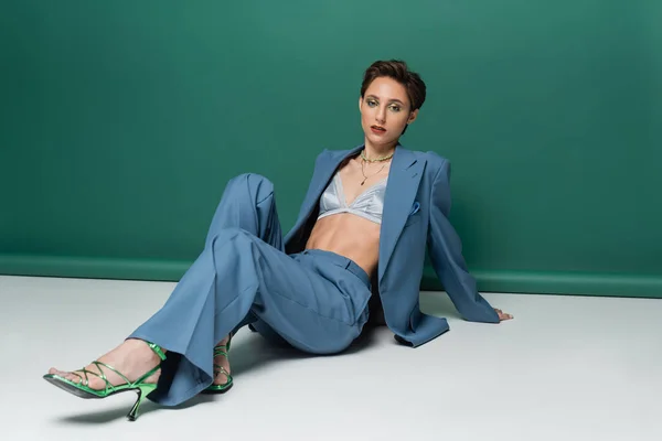 Full length of young woman with short hair posing in stylish suit with satin bra and heeled sandals on turquoise green — Stock Photo