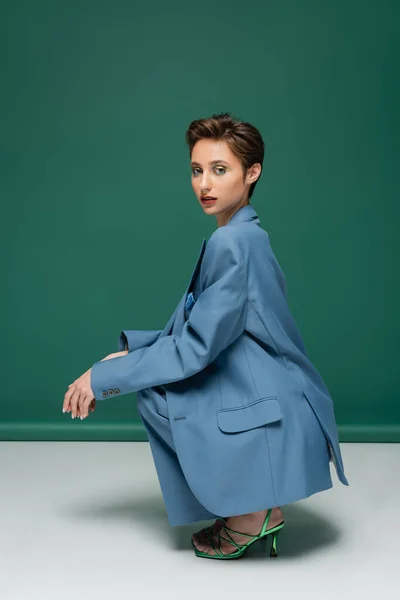 Young model with short hair sitting on haunches while posing in stylish pantsuit and heeled sandals on turquoise white background — Stock Photo
