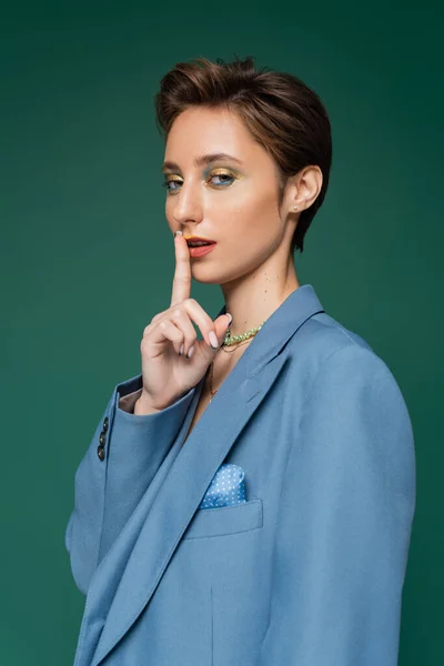 Молодая модель с короткими волосами, показывающая знак молчания, позируя в голубом блейзере, изолированном на бирюзовом — стоковое фото