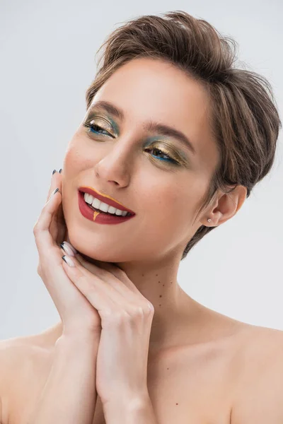 Jeune femme heureuse avec les cheveux courts et le maquillage lumineux regardant loin isolé sur gris — Photo de stock