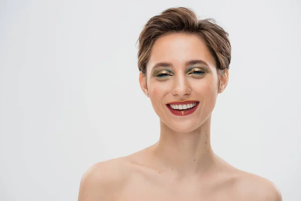 Überglückliche junge Frau mit kurzen Haaren und hellem Make-up blickt vereinzelt in die Kamera auf grau — Stockfoto