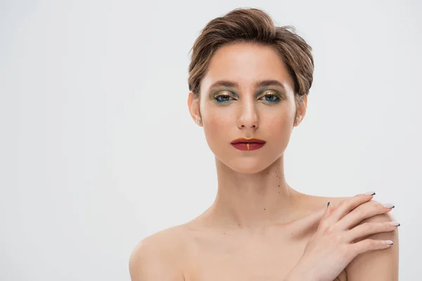 Портрет молодої жінки з яскравим макіяжем, який торкається голого плеча і дивиться на камеру ізольовано на сірому — Stock Photo