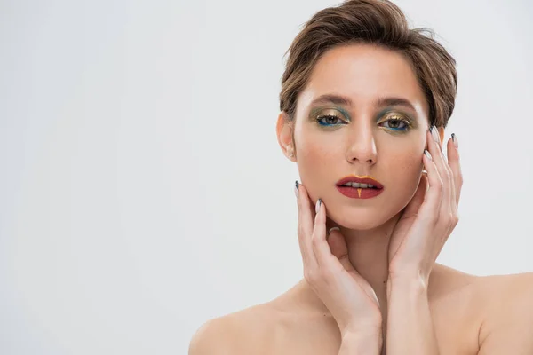 Retrato de modelo jovem com maquiagem brilhante tocando bochechas e olhando para a câmera isolada em cinza — Fotografia de Stock