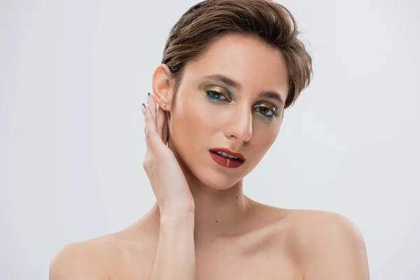 Retrato de modelo jovem com maquiagem brilhante fixando cabelo camisa e olhando para a câmera isolada em cinza — Fotografia de Stock